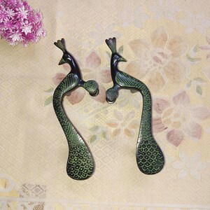 Może przedstawiać: Para drzwi w kształcie pawia wykonanych z mosiądzu z zielono-czarnym wykończeniem. Klamki są bogato zdobione piórami pawia i czubem na głowie.