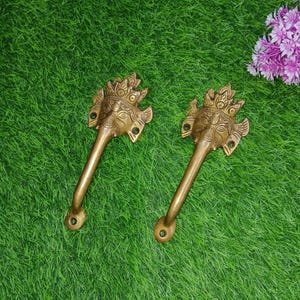 May include: Two gold-toned door handles with ornate designs featuring a face with a crown and a long nose. The handles are on a green grassy surface.