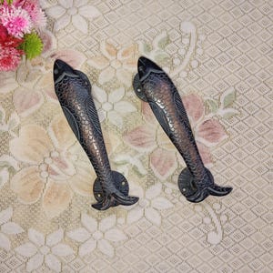 Op de afbeelding: Twee zwarte metalen deurgrepen in de vorm van een vis met een gedetailleerd ontwerp. De grepen bevinden zich op een lichtbeige achtergrond.