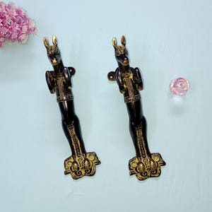 Puede incluir: Dos manijas de puerta decorativas con acabado negro y detalles dorados. Las manijas tienen forma de animales estilizados con cuerpos largos y bases ornamentadas. Una flor rosa y un pequeño objeto redondo e iridiscente también están en la imagen.