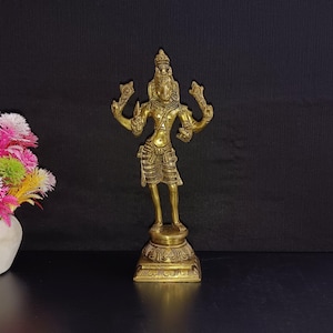 Puede incluir: Una estatua de bronce de color dorado que representa a una deidad hindú con cuatro brazos, de pie sobre una base cuadrada con tallas intrincadas. La estatua mide aproximadamente 25 cm de altura.