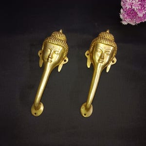 Peut inclure: Deux poignées de porte dorées avec un motif de visage de Bouddha. Les poignées sont longues et courbées, avec une base arrondie.
