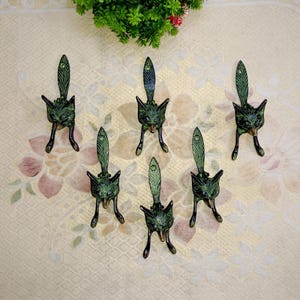 Può includere: Sette oggetti decorativi a forma di gatto, verdi e bronzo, sono disposti su una superficie a motivi floreali. Ogni gatto ha un volto dettagliato, una lunga coda e tre zampe. Gli oggetti sono in metallo e hanno un aspetto vintage.