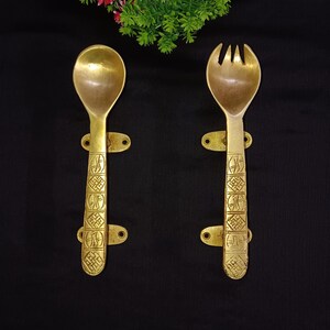 Puede incluir: Dos utensilios de servir de color latón: una cuchara y un tenedor. Ambos tienen mangos decorativos con un patrón grabado y orificios de montaje. Los utensilios se muestran sobre un fondo negro, con un arreglo floral verde y rojo en la parte superior.