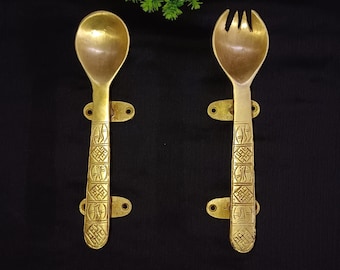 May include: Two brass-coloured serving utensils: a spoon and a fork. Both have decorative handles with an engraved pattern and mounting holes. The utensils are displayed against a black background, with a green and red floral arrangement at the top.