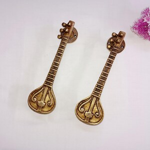May include: Two gold-toned metal door handles shaped like sitar musical instruments. The handles have intricate details and are mounted on a white background.