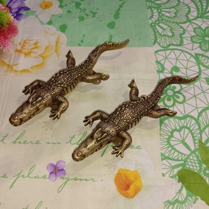 Op de afbeelding: Twee gouden alligator wandhaken. De alligators kijken in tegengestelde richtingen en hebben een getextureerd, schubbig oppervlak.