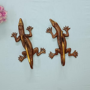 May include: Two bronze-colored lizard-shaped decorative handles. The lizards are detailed with textured scales and outstretched legs. The handles are mounted on a light blue surface, with a cluster of pink flowers in the upper left corner.