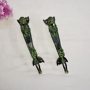 May include: Two antique door handles with a green and black finish. Each handle features a detailed face at the top and a decorative base. The handles are made of metal and have a vintage aesthetic.