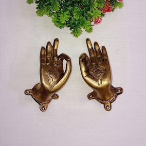 May include: Two brass-colored hand sculptures in the 'okay' gesture, mounted on small bases. Each hand features intricate detailing and a diamond-shaped design on the palm. The hands are positioned against a white background, with greenery in the upper portion.
