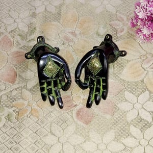 Könnte beinhalten: Ein Paar schwarze Metall-Türgriffe in Form von Händen mit grünen Akzenten und goldenen Details. Die Griffe haben ein rautenförmiges Design in der Mitte.