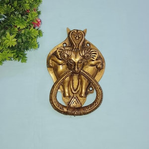 May include: A brass door knocker featuring an intricate design of a mythical creature with horns and a human face. The knocker is shaped like a ring with an ornate design.