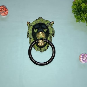 以下が含まれることがあります： 黒と緑のライオンの頭のドアノッカーで、金色のリングが付いています。ノッカーは薄い青色の壁に取り付けられています。