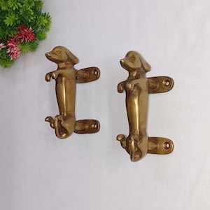 Può includere: Un paio di maniglie per porte in ottone a forma di bassotto. Le maniglie sono in posizione eretta con la testa girata di lato. Le maniglie sono montate su uno sfondo bianco.