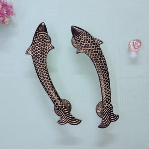 May include: A pair of black metal door handles shaped like fish with a silver-coloured design. The handles are curved and have a detailed fish scale pattern.