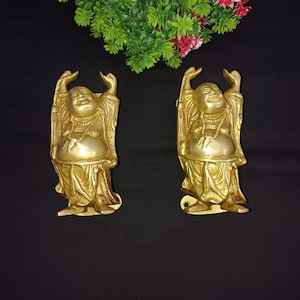 May include: Two gold-coloured metal figurines of a laughing Buddha, each with raised arms and a round belly. The figurines are standing on a flat base.