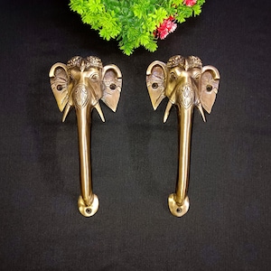 May include: A pair of brass door handles shaped like elephant heads with intricate details. The handles are a golden colour and have a long, curved design.