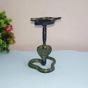 May include: A black metal incense burner shaped like a cobra with a green and black patterned body. The burner has a round top with a raised design.