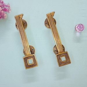 May include: Two antique brass door handles with a square design and a textured pattern. The handles are rectangular with a decorative top and bottom. A pink glass decorative object and pink flowers are in the background.