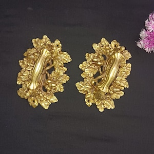 Może przedstawiać: Dwa złote metalowe uchwyty do drzwi w kształcie liści dębu z żołędziami. Uchwyty mają ozdobny wzór i nadają się do szafki lub szuflady.