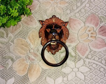 May include: A brown lion's head door knocker with a black ring. The knocker is on a white background with a floral pattern.