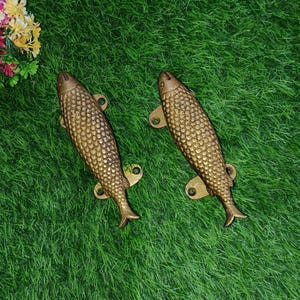 May include: Two gold-toned fish-shaped door handles with detailed scales. The handles are on a green artificial grass background.