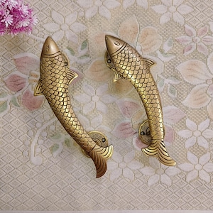 Pode incluir: Duas maçanetas de porta em forma de peixe douradas com escamas detalhadas e um design curvo.