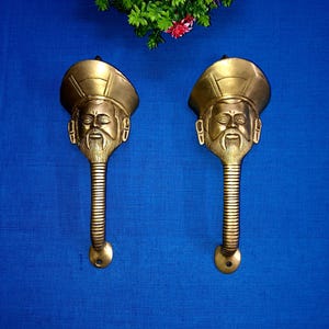 May include: Two antique brass door handles with a face design. The handles are in the shape of a person's head with a hat and a beard. The handles are on a blue background.