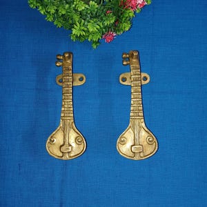Peut inclure: Une paire de poignées de porte en métal doré en forme d'instruments de musique sitar. Les poignées sont montées sur un fond bleu.