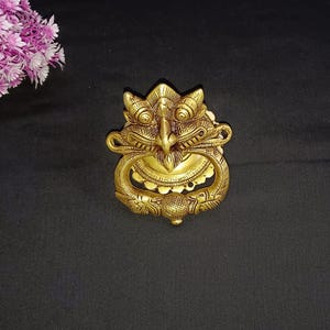 Può includere: Un batacchio per porta in metallo dorato a forma di creatura mitologica con un'espressione feroce. Il batacchio presenta dettagli intricati e un anello per tirare.