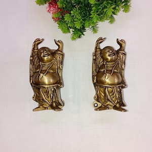 May include: Two gold-coloured metal figurines of the laughing Buddha, each with raised arms and a joyful expression. The figurines are standing on a white surface.