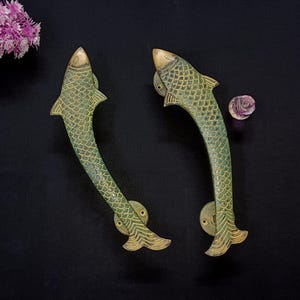 May include: Two antique brass door handles shaped like fish. The handles have a green patina with gold accents, set against a black background. The fish scales are detailed. Each handle is approximately 20 cm long.