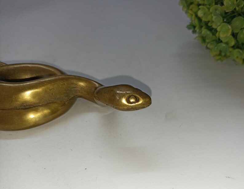 May include: A brass snake figurine with a detailed, textured surface. The snake is coiled, with its head in the foreground, showcasing a detailed eye. The figurine has a polished, golden finish, and is set against a white background.