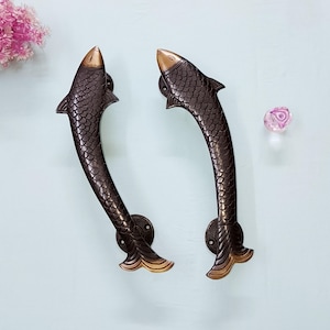 May include: Two curved door handles shaped like fish, featuring a dark bronze-toned finish with detailed scale textures and lighter metallic accents on the heads and tails. Each handle includes a circular mounting plate at the base for installation.