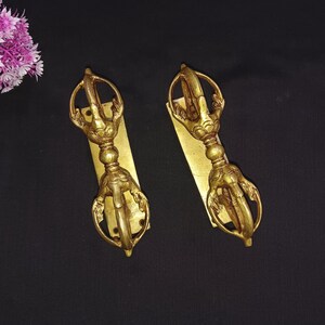 May include: Two gold-colored metal door handles with intricate designs. Each handle features a circular ring with a decorative knot and a long, curved arm with a pointed tip. The handles are mounted on rectangular plates.
