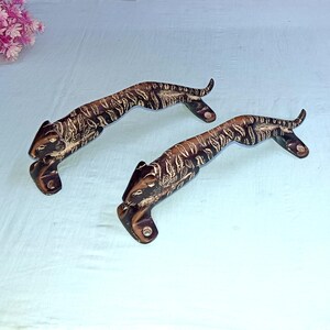 Peut inclure: Deux poignées de porte décoratives en forme de tigres. Les poignées sont marron avec des détails noirs et blancs, posées sur une surface bleu clair. Elles sont en métal et ont un aspect vintage. Mesurent environ 20 cm de long.