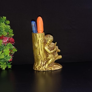 Puede incluir: Un portaplumas de metal dorado con forma de tronco de árbol con un querubín sentado en una rama. El querubín sostiene un pequeño pájaro en sus manos.