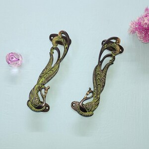 Op de afbeelding: Twee sierlijke messing deurgrepen in de vorm van pauwen met groene en gouden accenten.