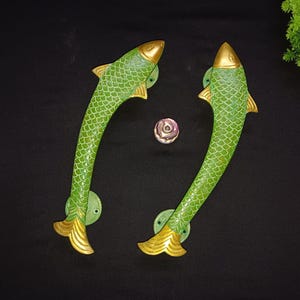 May include: Two green fish-shaped door handles with gold detailing. The handles feature a textured scale pattern and are mounted on round, green bases. A small, decorative knob is centered between them.