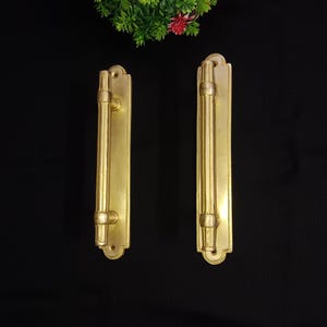 May include: Two gold-toned door handles with a rectangular shape and a long, cylindrical pull. The handles have a decorative design around the edges.