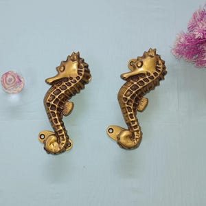 Könnte beinhalten: Zwei messingfarbene Schrankgriffe in Form von Seepferdchen. Die Seepferdchen haben strukturierte Körper und Flossen und sind auf einer hellblauen Oberfläche montiert. Eine kleine, dekorative Glaskugel und rosa Blumen sind ebenfalls zu sehen.