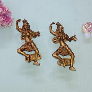 Op de afbeelding: Twee antieke messing deurklinken, elk met een dansende figuur in een dynamische pose. De figuren zijn gedetailleerd met kleding en sieraden. De deurklinken zijn gemonteerd op rechthoekige platen met schroefgaten. Een roze bloemstuk en een decoratieve glazen bol zijn op de achtergrond.