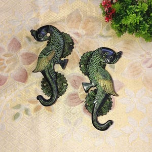 May include: Two green and gold metal seahorse door handles with intricate details. The handles are mounted on a floral design.