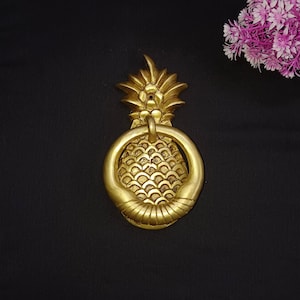 May include: A gold-colored pineapple door knocker. The knocker features a detailed pineapple design with a ring for knocking. The pineapple is made of metal and has a textured surface. The door knocker is mounted on a black surface.