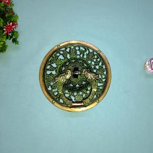 May include: An antique brass door knocker with a green patina, featuring a detailed floral and dragon design. The round knocker is set against a light teal background.
