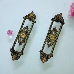 Puede incluir: Dos tiradores decorativos de color bronce con un diseño floral. Los tiradores tienen forma rectangular con detalles ornamentados, incluyendo una flor central y detalles de hojas. Los tiradores están pintados con tonos verdes y marrones.