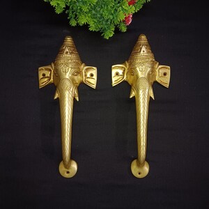 Könnte beinhalten: Zwei goldfarbene Türklinken in Form eines Elefantenkopfes mit aufwendigen Details. Die Griffe sind auf einem schwarzen Hintergrund montiert.