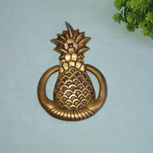 Op de afbeelding: Een gouden ananas deurklopper met een ringvormige handgreep. De ananas is gedetailleerd met een getextureerd oppervlak en een gesneden ontwerp.