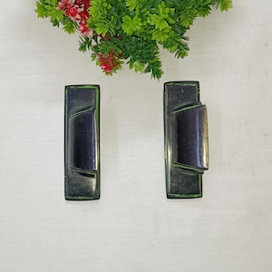 May include: A pair of vintage metal cabinet pulls. Each pull features a rectangular base with a dark, curved central section and a gold-toned accent. The pulls are dark green with a distressed finish. Decorative greenery with red flowers is in the background.