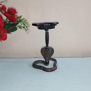 以下が含まれることがあります： 丸いトップを持つ黒い金属製の蛇の形をしたお香立て。お香立ては薄い青色の表面の上に置かれています